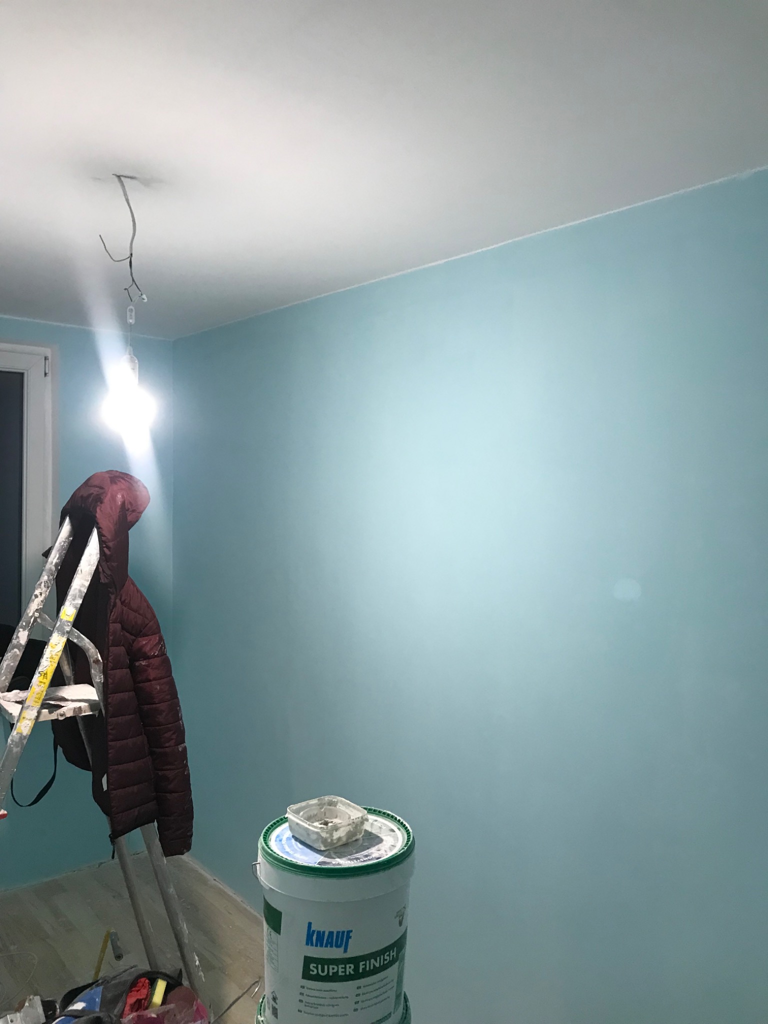 Świeżo pomalowane na błękitno ściany w pokoju, widoczna drabina oparta o ścianę, wiadro z gładzią Knauf Super Finish, wisząca żarówka bez klosza.