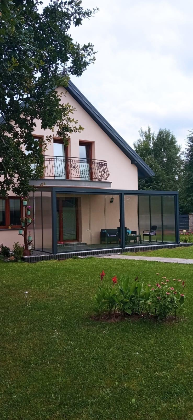 Nowoczesna szklana zabudowa tarasu przylegająca do domu z balkonem, widok na zielony trawnik z kwiatami.