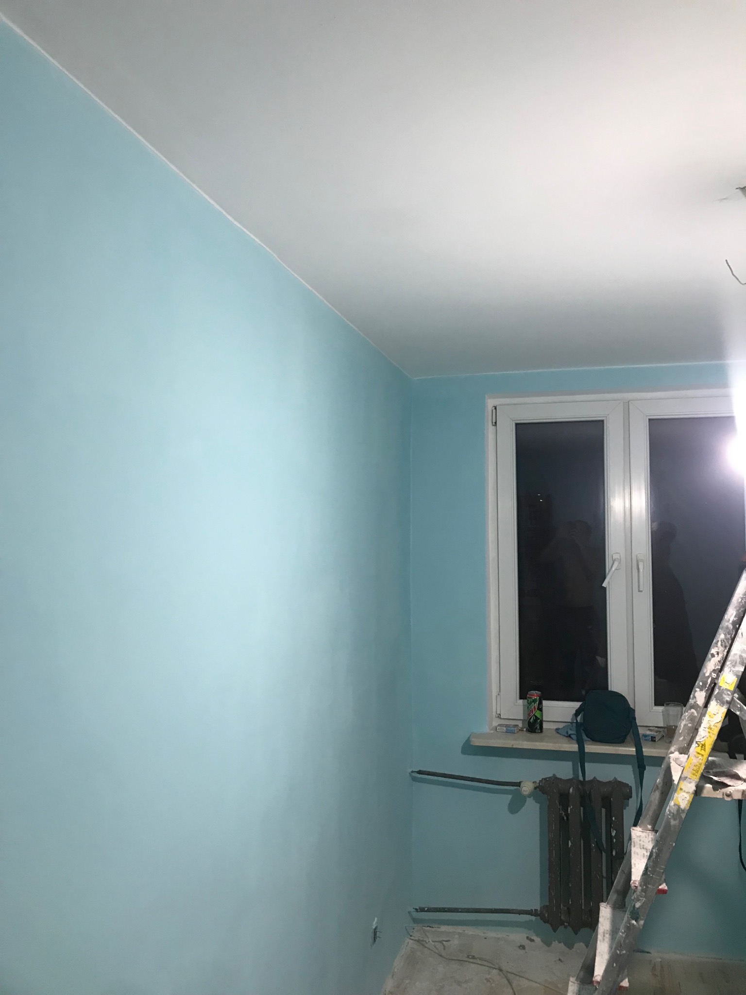 Świeżo pomalowane na błękitno ściany pokoju z grzejnikiem pod oknem; widoczna drabina malarska, puszka napoju i torba na parapecie.