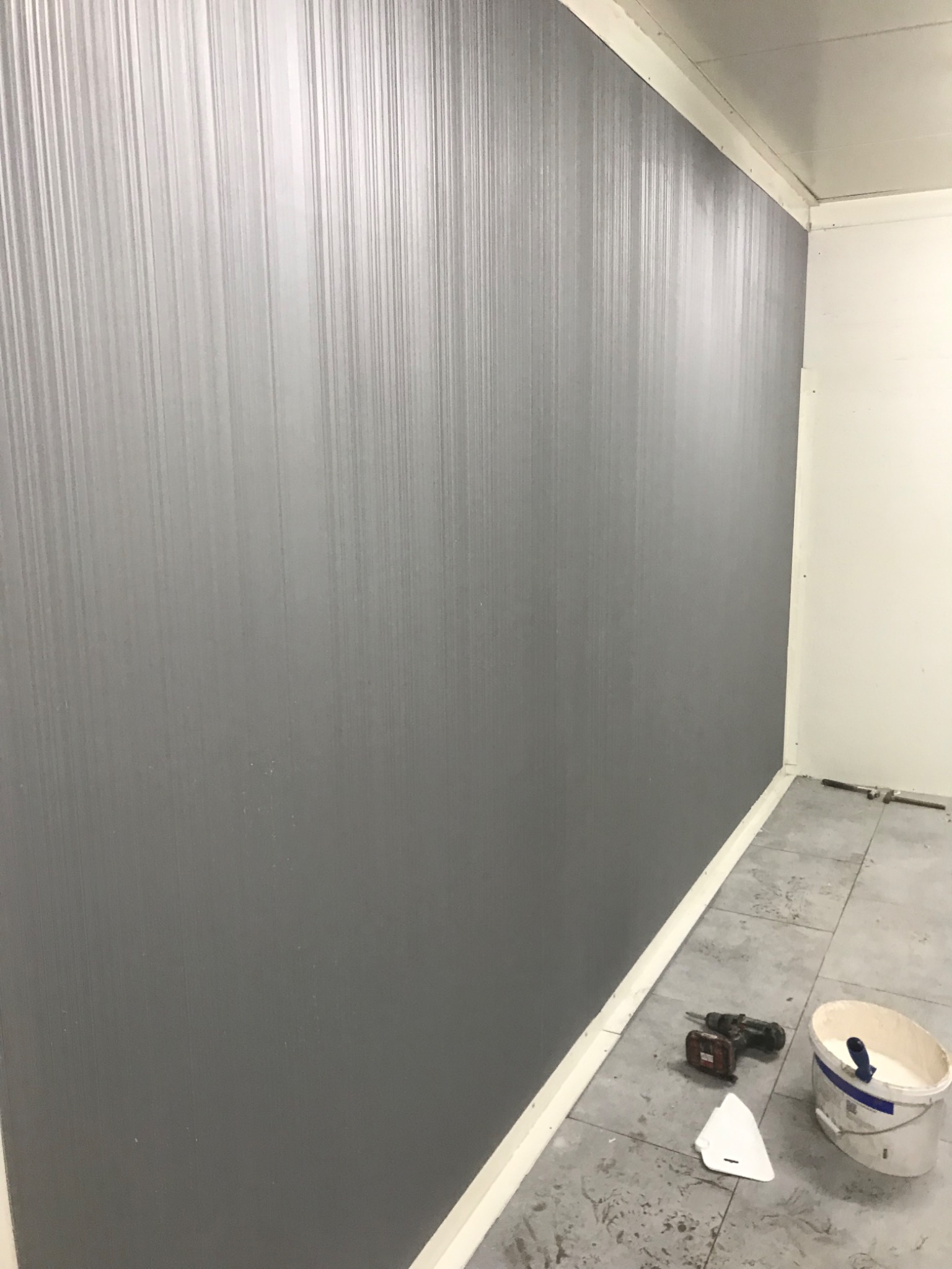 Świeżo pomalowana szara ściana z pionowymi smugami, na podłodze wiadro z farbą, szpachelka i wiertarko-wkrętarka.