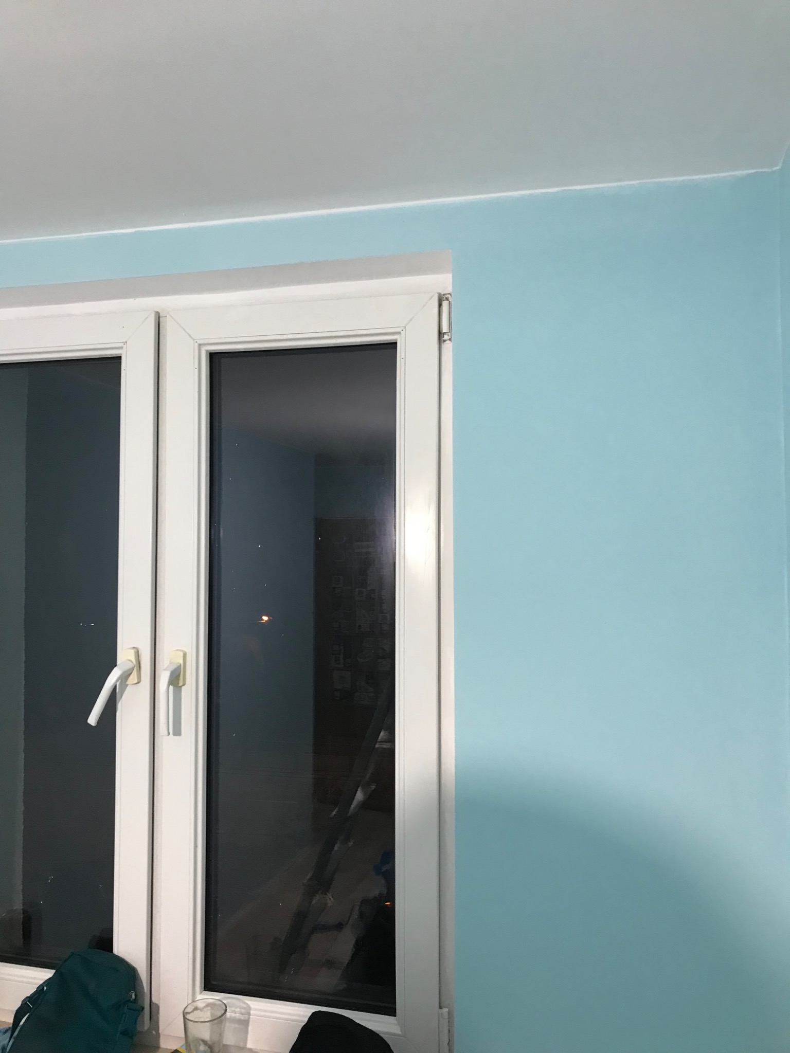 Świeżo pomalowana na błękitno ściana w pokoju, widoczna biała rama okienna z klamkami, na parapecie torba i szklanka, w odbiciu okna widoczna drabina.