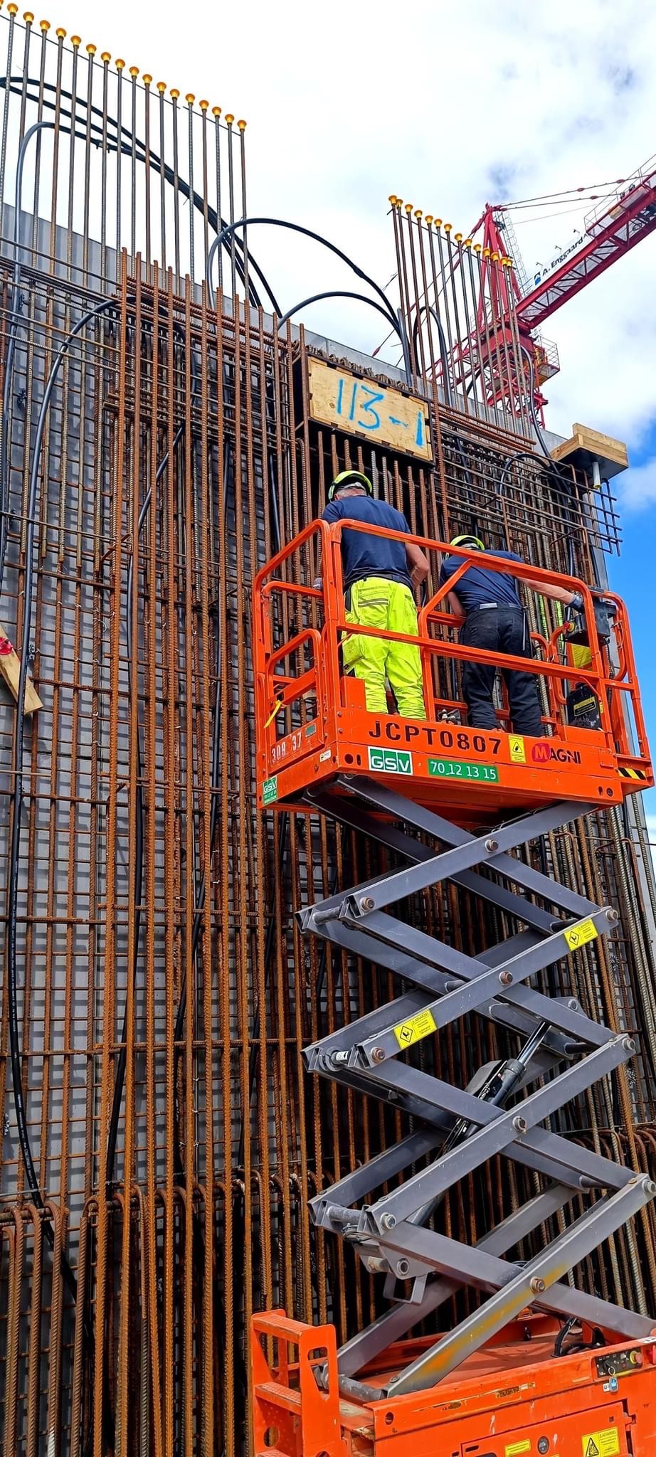 Dwóch pracowników w żółtych i granatowych ubraniach roboczych na podnośniku koszowym, montujących zbrojenie ściany z prętów stalowych, widoczna tabliczka z numerem '113-1' i dźwig budowlany w tle.