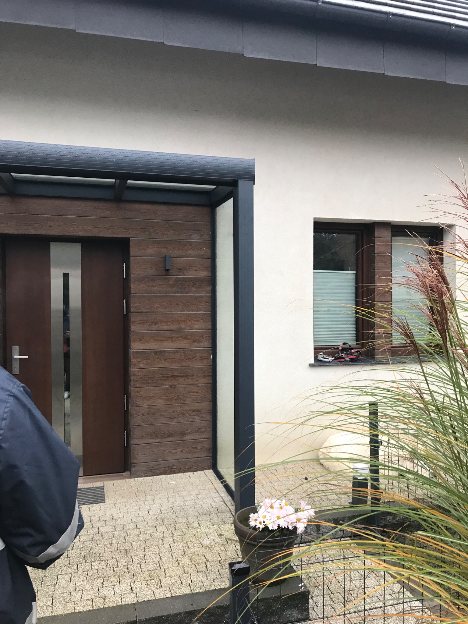 Elewacja domu jednorodzinnego z drewnianą okładziną, nowoczesne drzwi wejściowe z metalowym akcentem, okno z żaluzjami plisowanymi i dekoracją, donica z kwiatami przy wejściu.