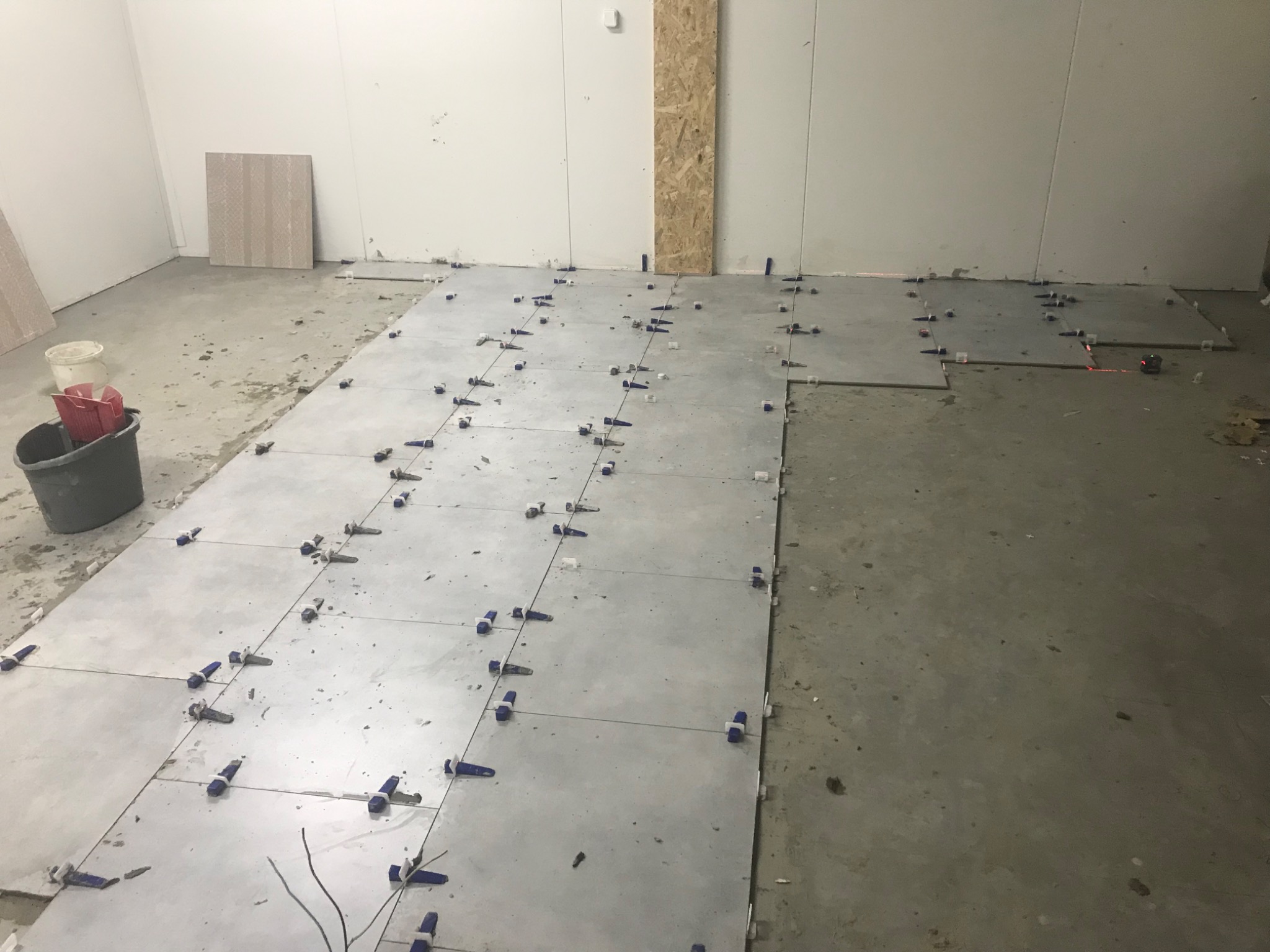 Układanie szarych płytek podłogowych z użyciem niebieskich klinów poziomujących na nierównym, betonowym podłożu. W tle widoczne wiadro i płyty opierające się o ścianę.