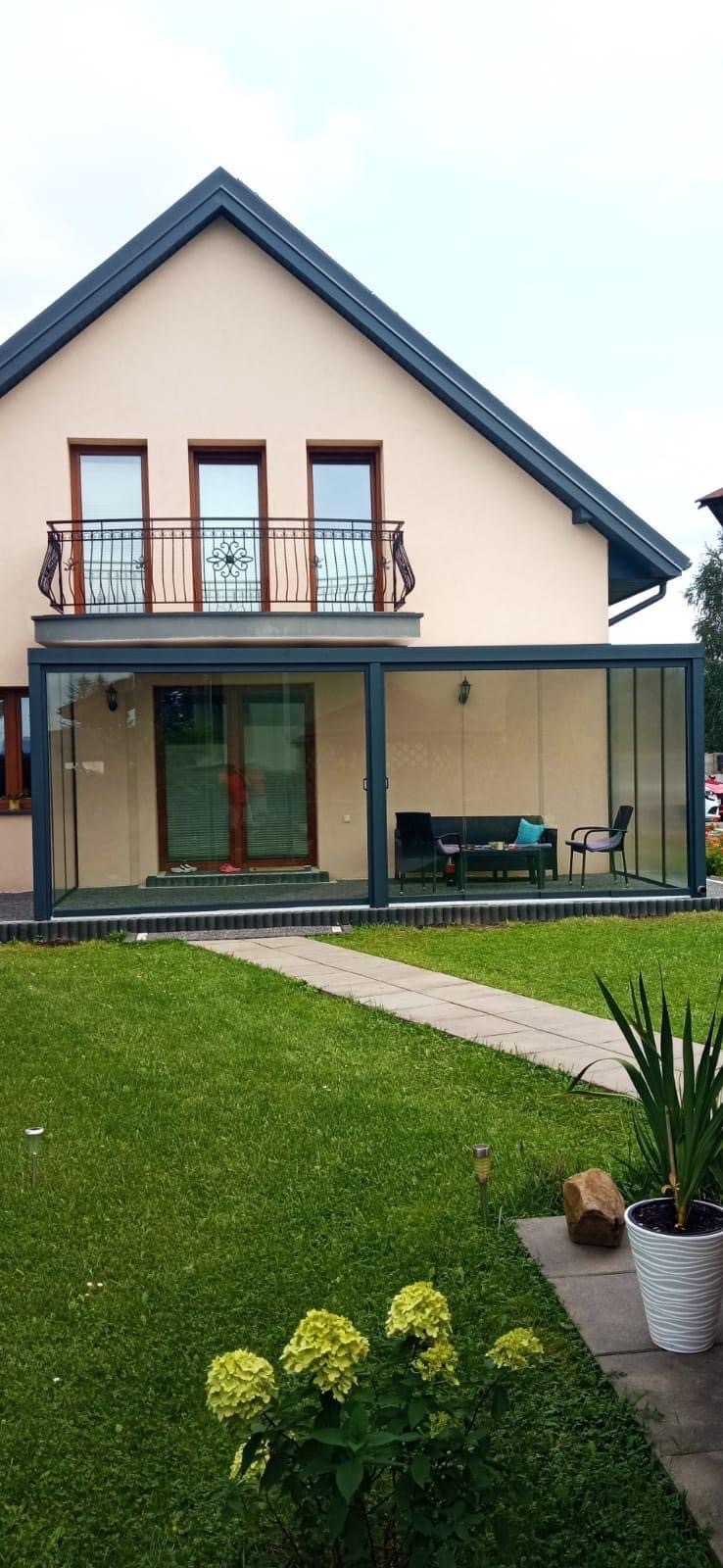 Szklana zabudowa tarasu przy domu z balkonem z kutą balustradą, widok z ogrodu na meble wypoczynkowe wewnątrz.