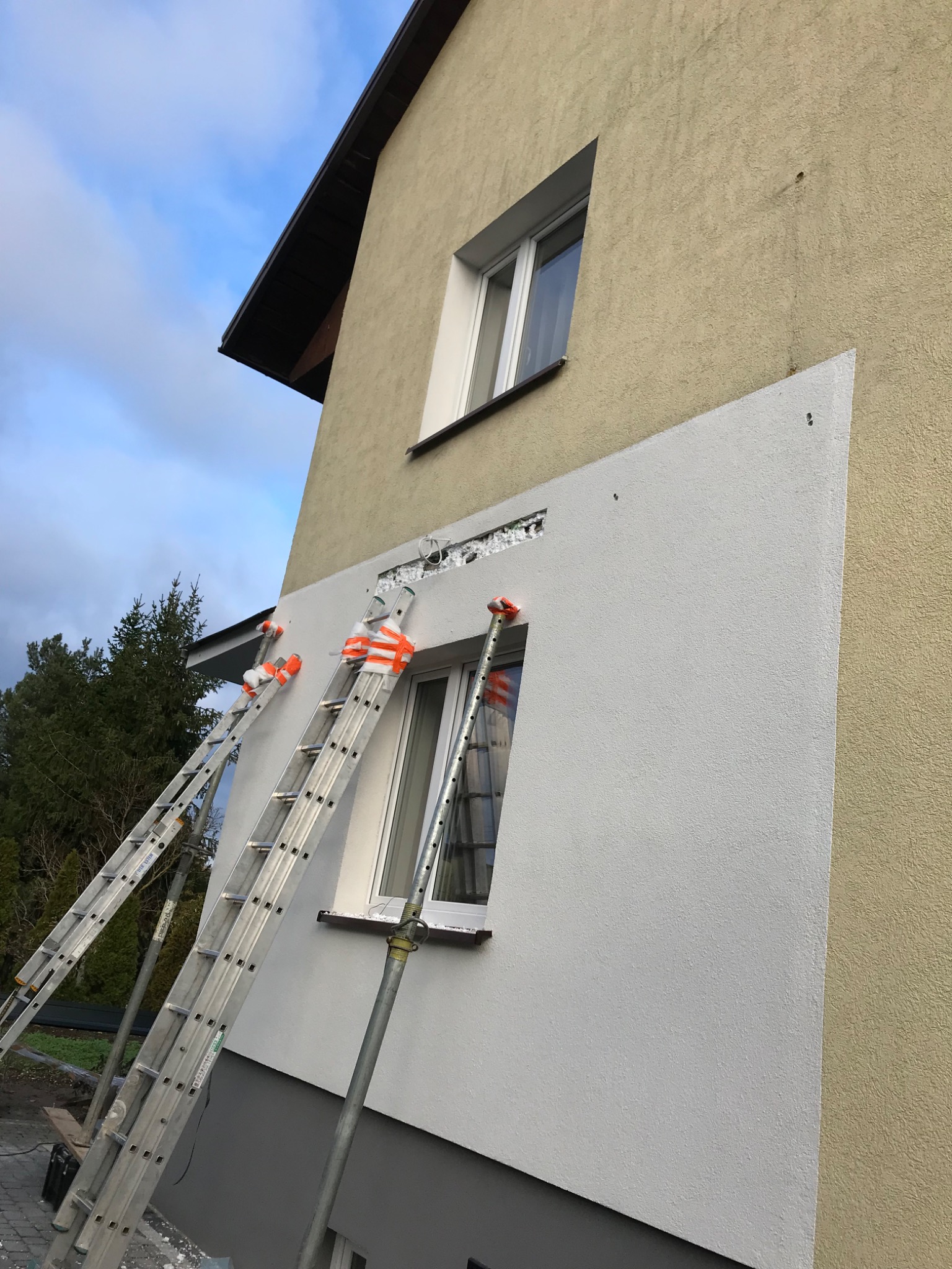 Ocieplanie ściany budynku styropianem, widoczne dwie drabiny opierające się o ścianę, fragment otynkowanej powierzchni i okno