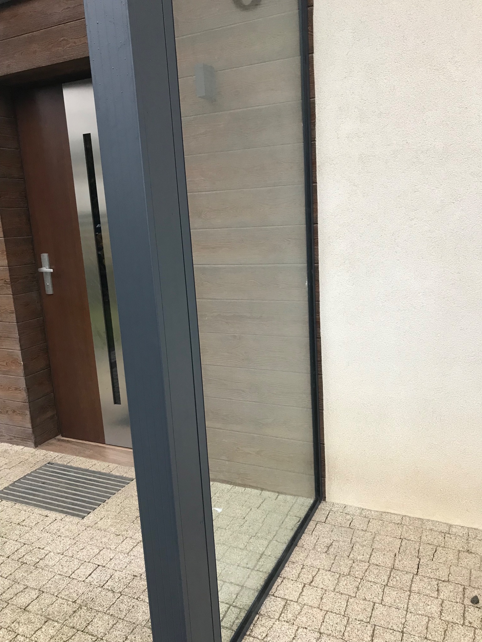 Nowoczesna elewacja budynku z drewnianą stolarką okienną i drzwiami wejściowymi, widoczna kostka brukowa na podłożu.