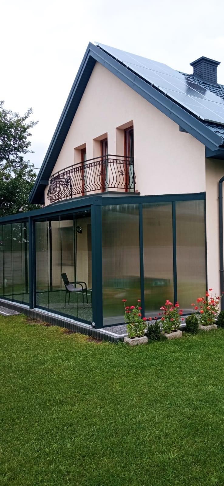 Szklana zabudowa tarasu przy domu jednorodzinnym z balkonem z kutą balustradą, widok z zewnątrz na krzesło wewnątrz zabudowy i rabatki z czerwonymi kwiatami.