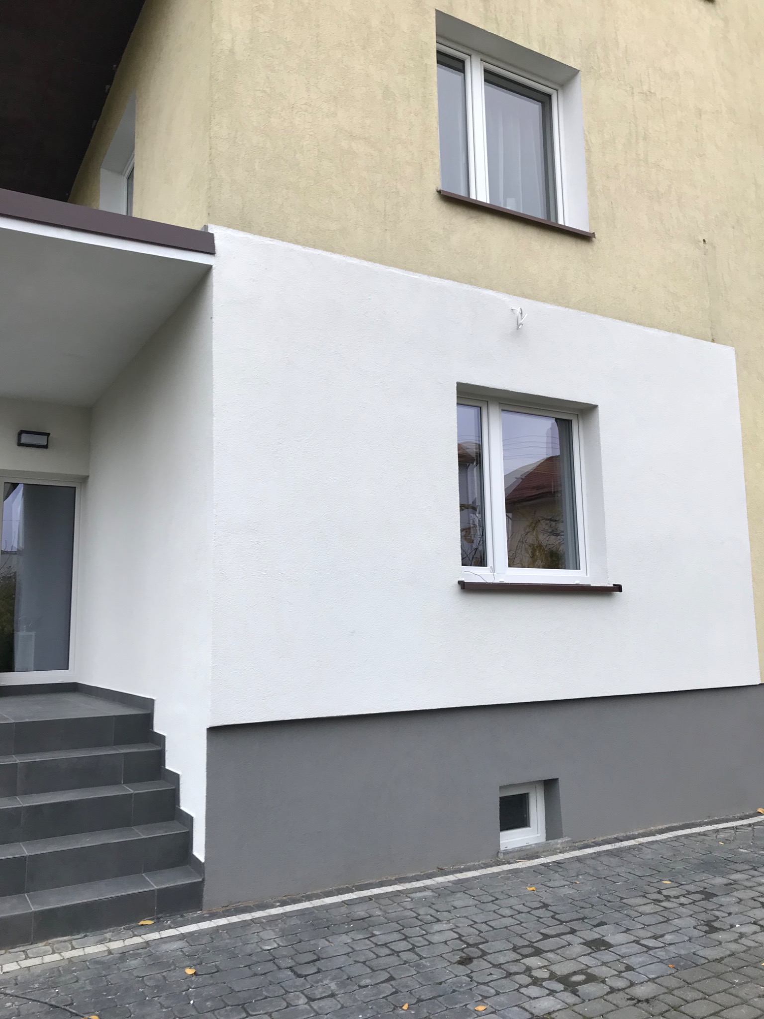 Odnowiona elewacja budynku z białym tynkiem, szarym cokołem i grafitowymi schodami wejściowymi wyłożonymi płytkami.