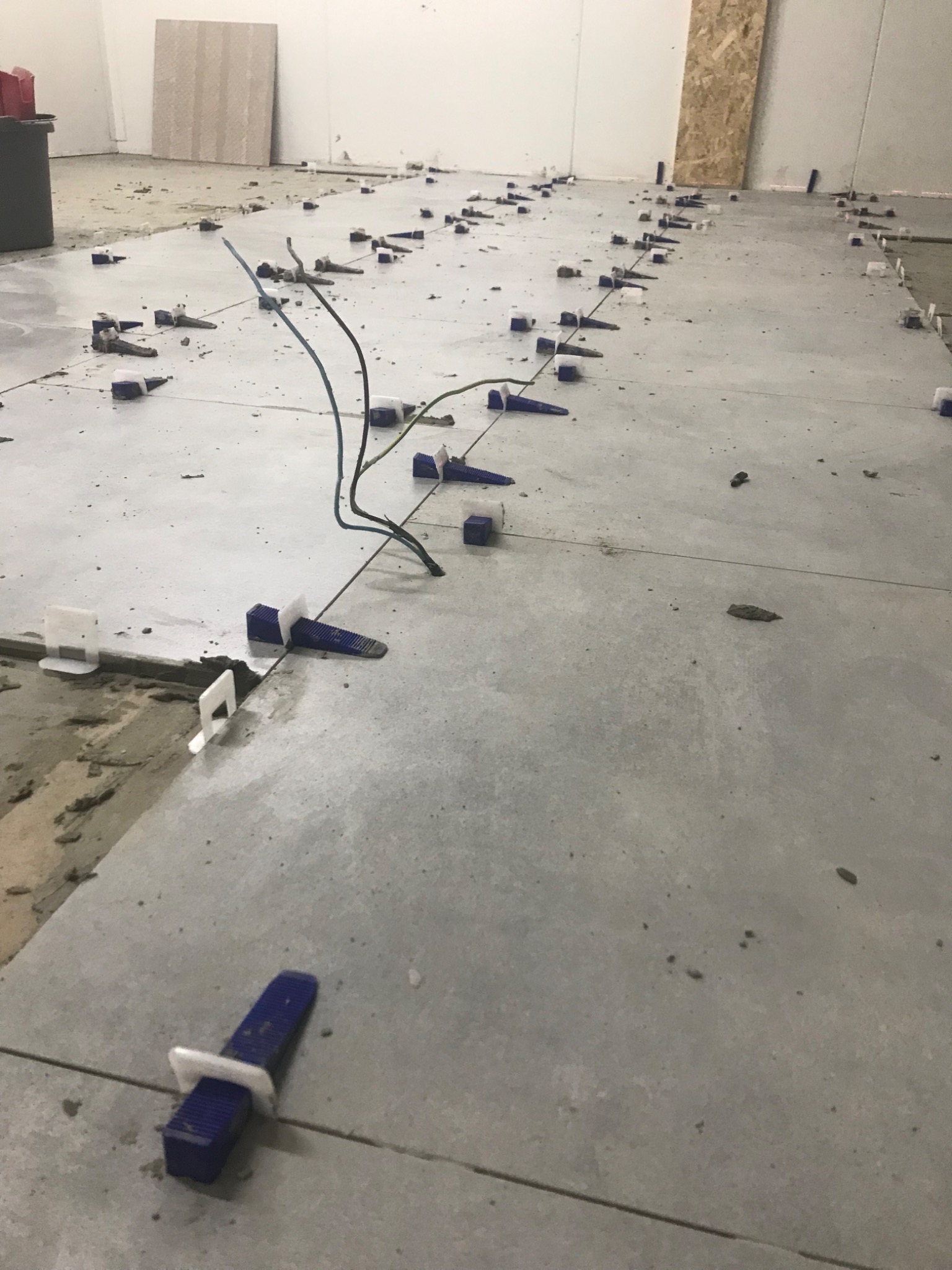 Układanie płytek podłogowych w trakcie realizacji, widoczne niebieskie kliny dystansowe i resztki zaprawy, wystające przewody elektryczne. Płytki w odcieniach szarości z widocznymi fugami.