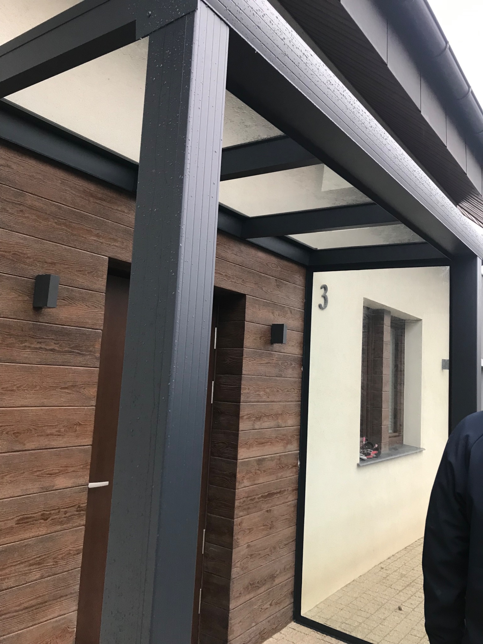 Nowoczesne zadaszenie nad wejściem do domu z drewnianą elewacją i numerem 3 na białej ścianie, widoczne krople deszczu na czarnej konstrukcji.