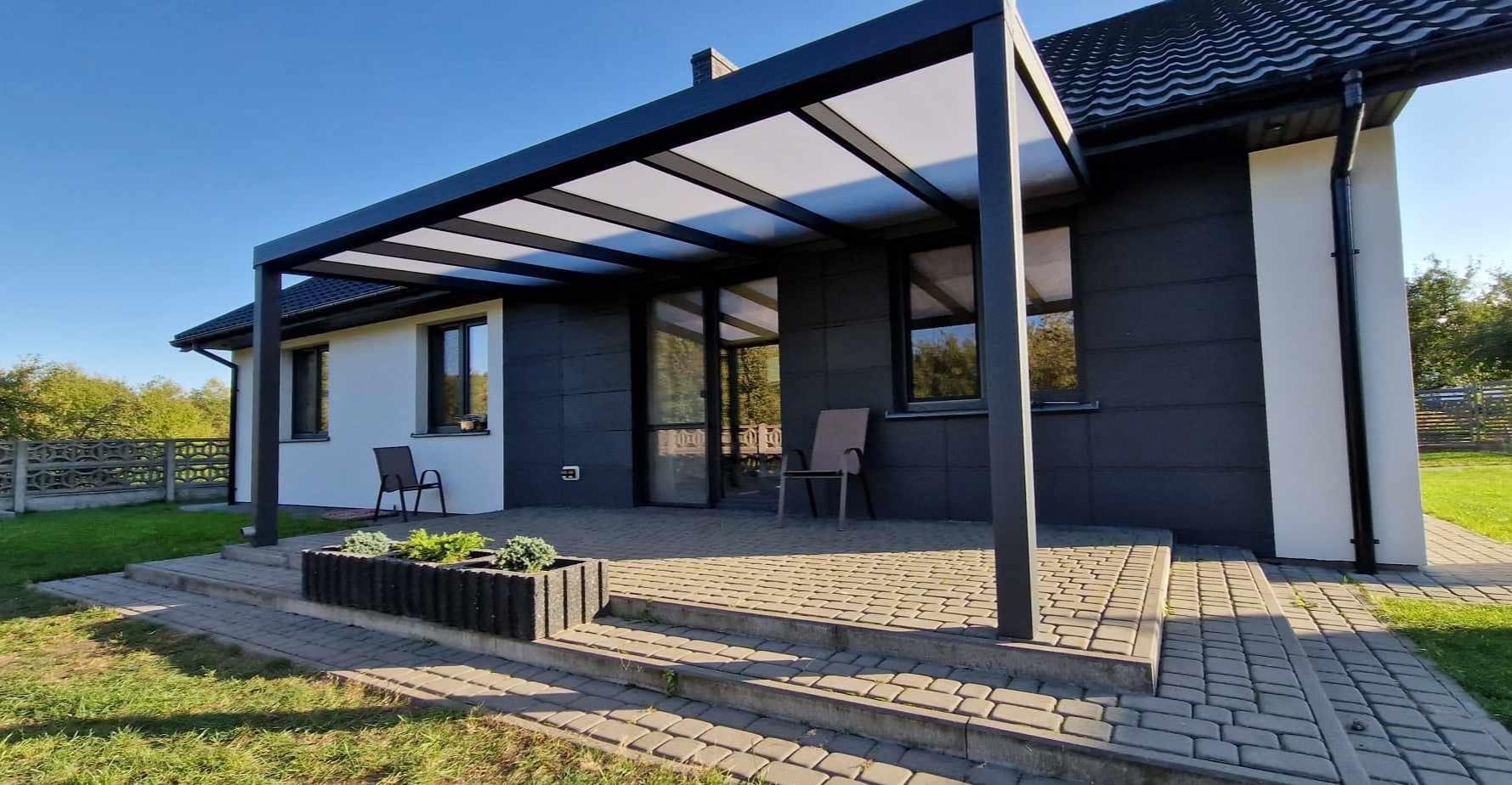 Nowoczesna pergola z czarną konstrukcją i przezroczystym zadaszeniem przylegająca do domu z szarą elewacją, na tarasie dwie szare krzesła i betonowa donica z zielonymi roślinami.