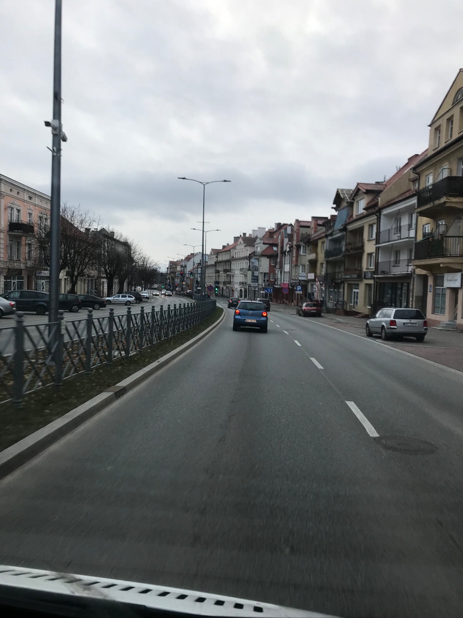 Ulica miejska z budynkami po obu stronach, widok z perspektywy pojazdu w ruchu, pochmurne niebo.