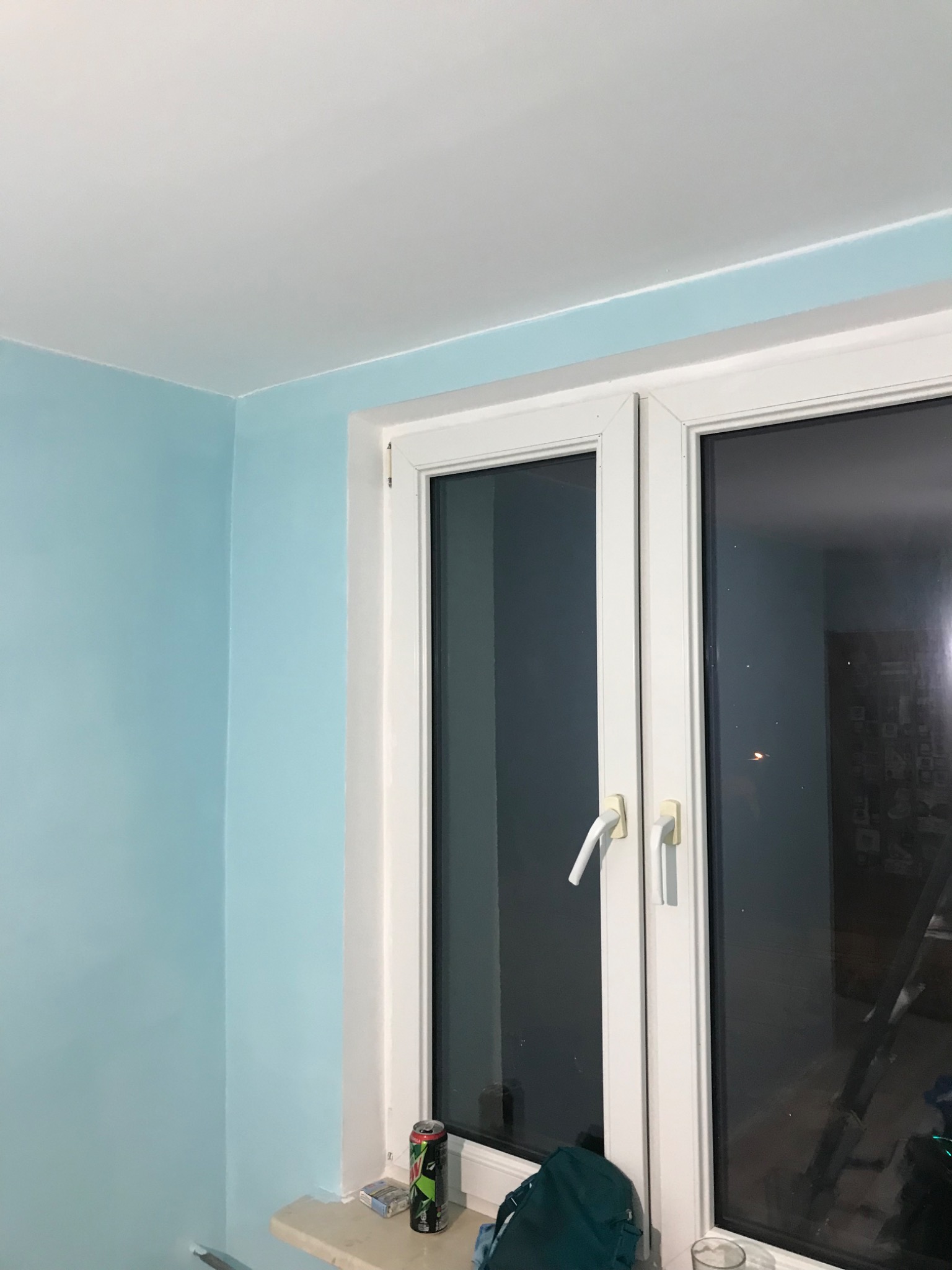 Świeżo pomalowany pokój w odcieniach błękitu, z białym sufitem i ramą okienną. Na parapecie puszka napoju i paczka papierosów. W szybie okna odbija się sylwetka drabiny.