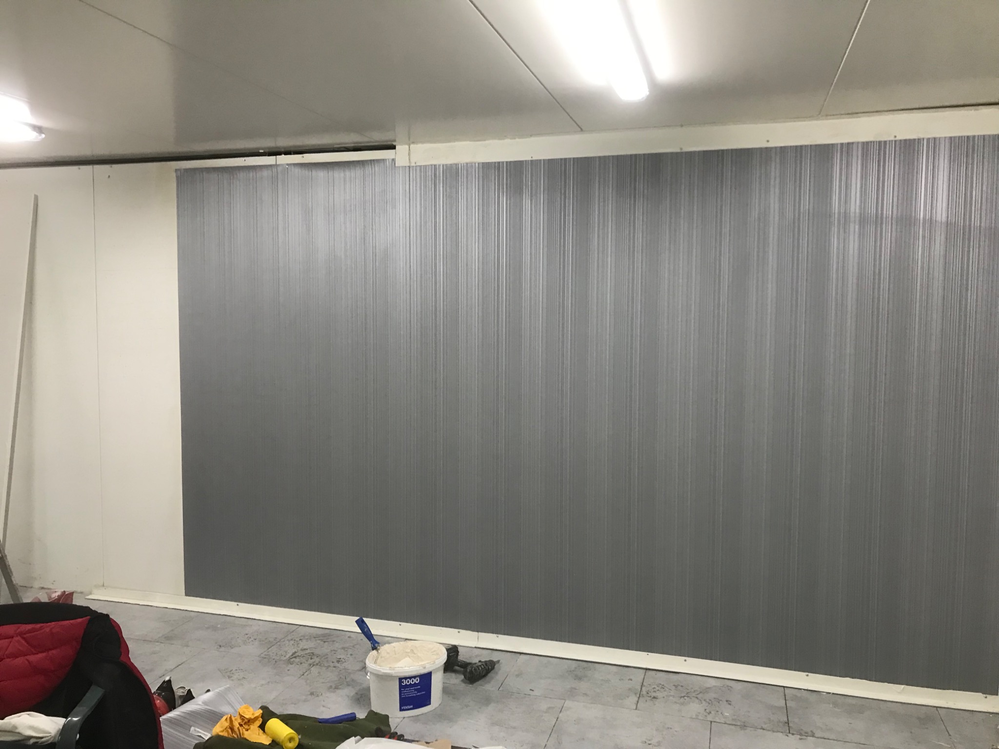 Szara ściana z pionowymi paskami w biurze lub lokalu usługowym, białe listwy przypodłogowe i sufit, wiadro z masą szpachlową i narzędzia na podłodze.