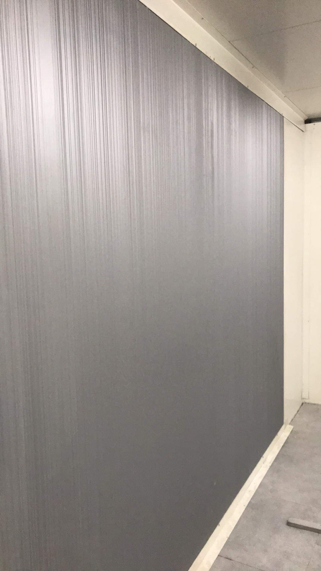 Świeżo pomalowana, szara ściana z pionowymi, delikatnymi smugami po wałku, widoczna krawędź sufitu i przypodłogowa listwa.