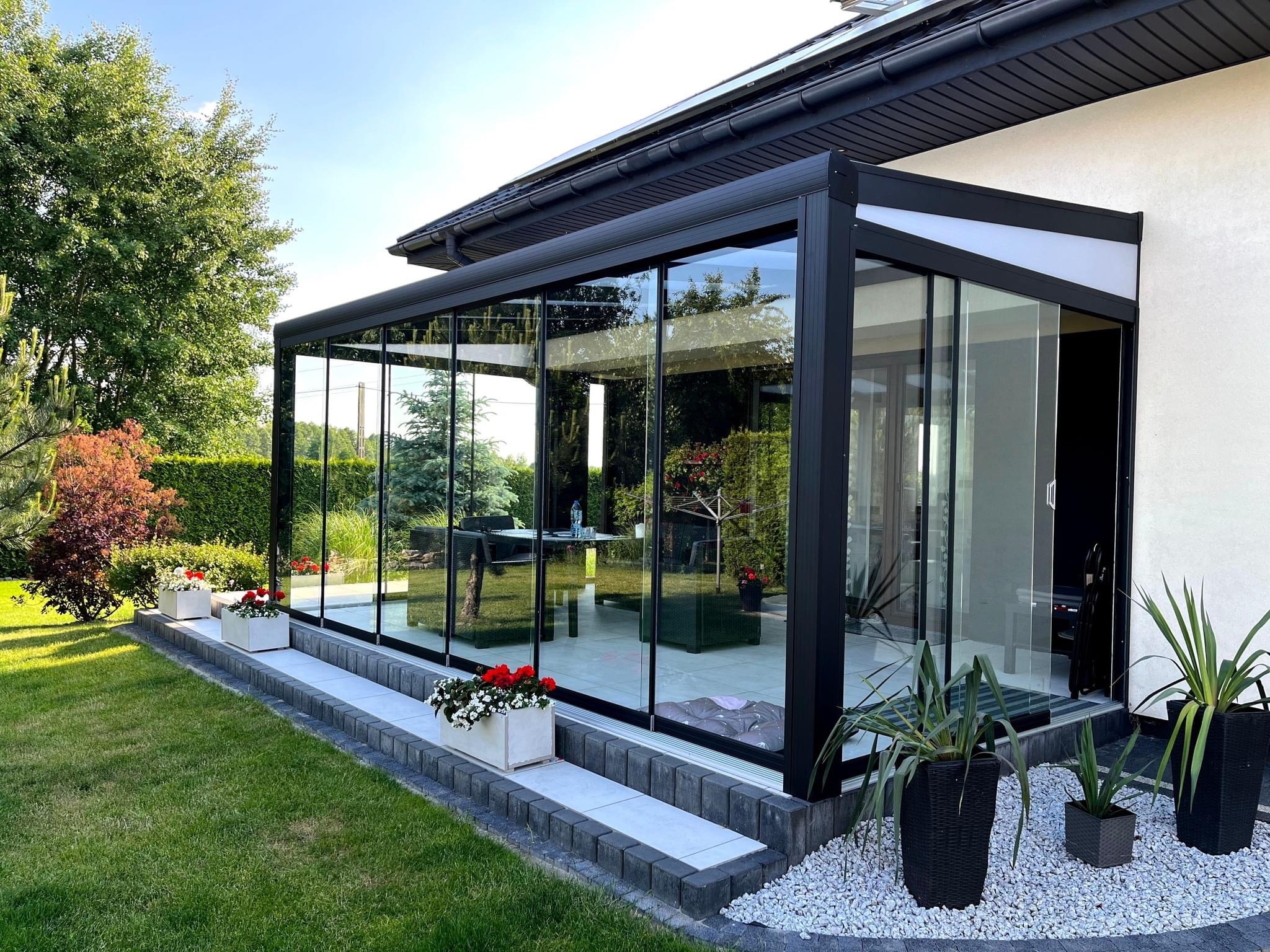 Nowoczesna, przeszklona weranda z czarną ramą, przylegająca do białego budynku, otoczona zielonym trawnikiem i ozdobnymi kwiatami w donicach.