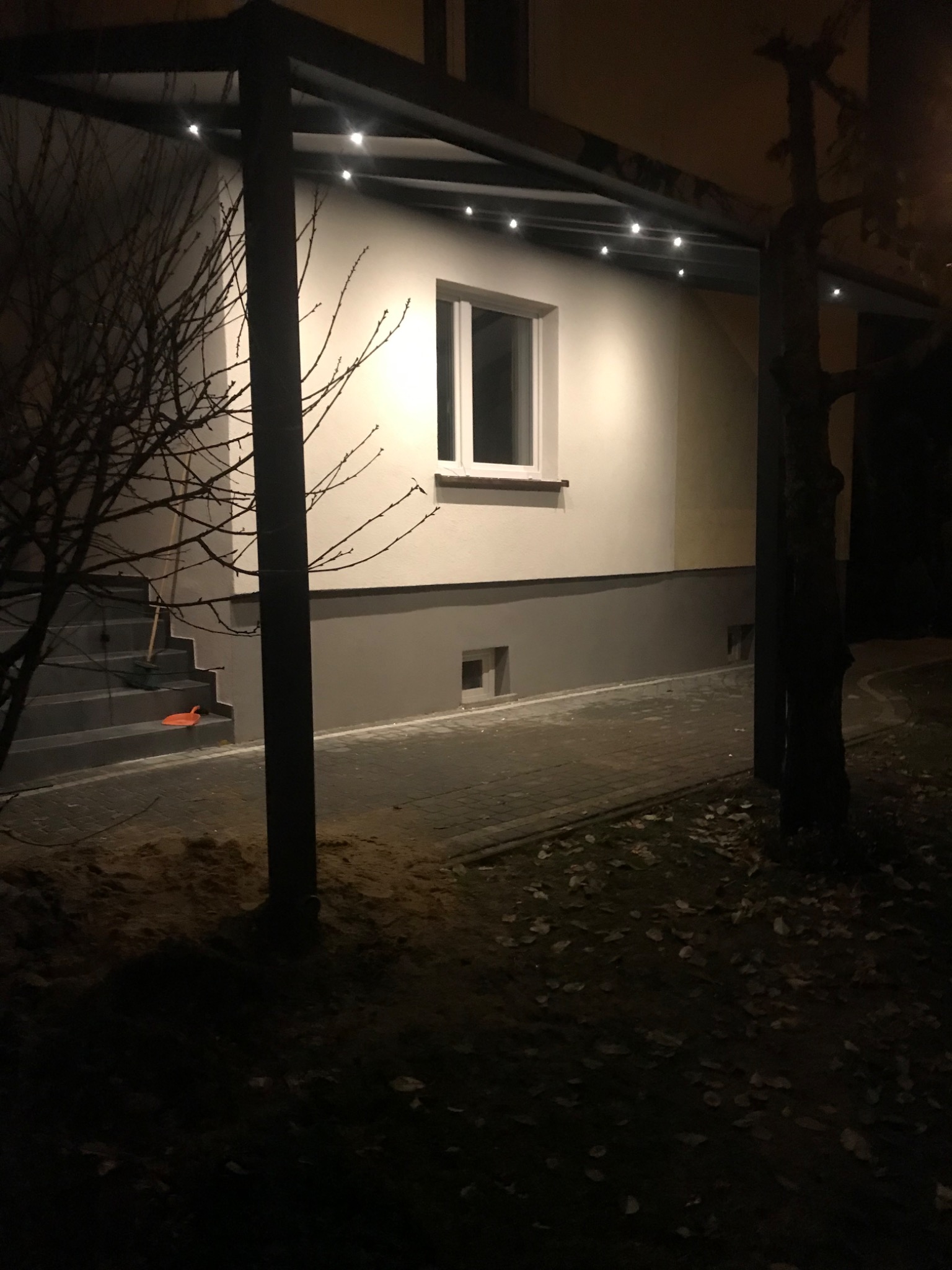 Świeżo pomalowana elewacja budynku z szarą podmurówką i oświetleniem LED pod okapem, widoczne schody wejściowe z pozostawioną szufelką i miotłą.