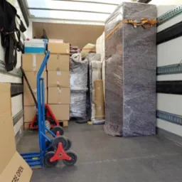 Wnętrze busa załadowane kartonami, meblami owiniętymi folią stretch i wózkiem transportowym z trójkołowymi gwiazdami na schody.