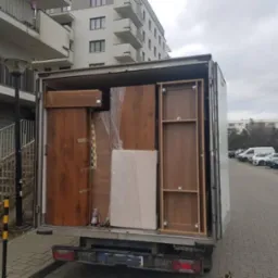 Otwarta paka samochodu dostawczego załadowana meblami, w tym szafa, komoda i materac, zabezpieczone folią, na tle bloku mieszkalnego i parkingu.