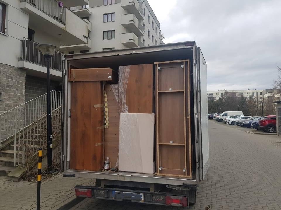 Otwarta paka samochodu dostawczego załadowana meblami, w tym szafa, komoda i materac, zabezpieczone folią, na tle bloku mieszkalnego i parkingu.