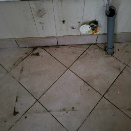 Szufeleczka Sp. z o. o. - Brudna podłoga z płytek i ściana z widocznymi zabrudzeniami, wystającą rurą kanalizacyjną i otwartą puszką elektryczną z wystającymi przewodami.