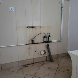 Szufeleczka Sp. z o. o. - Ściana z panelami, widoczne rury, zawory, przyłącza kanalizacyjne i elektryczne przed montażem umywalki, ślady po starej instalacji i zabrudzenia na ścianie i podłodze.