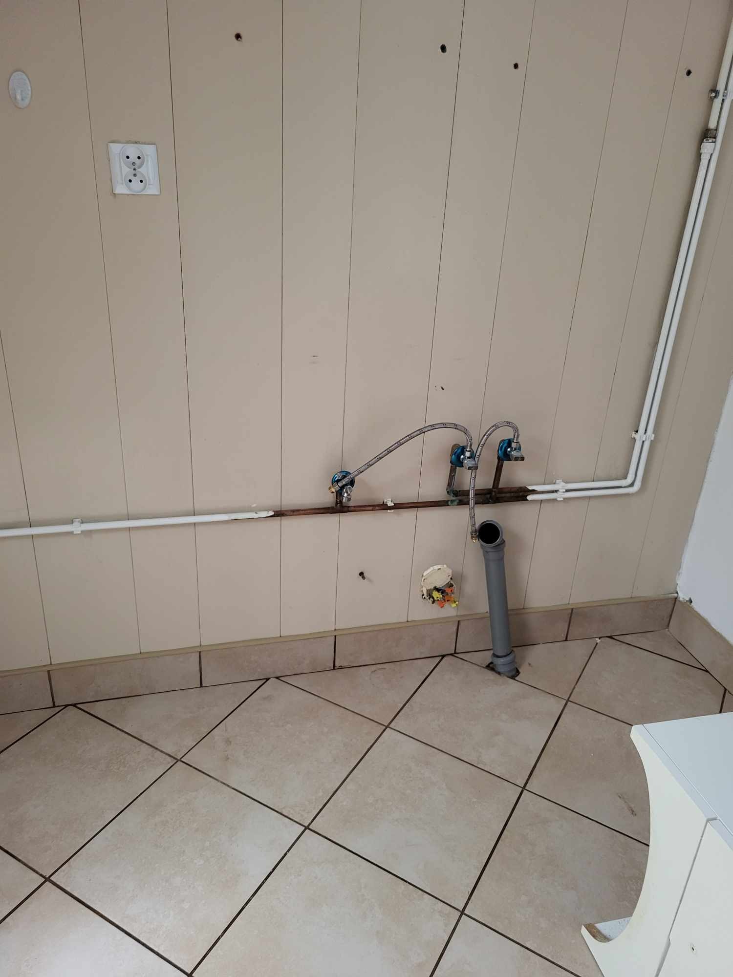 Fragment łazienki w trakcie remontu: widoczne przyłącza wodne, rury PCV, instalacja elektryczna, beżowe płytki na ścianach i podłodze z widocznymi śladami po pracach budowlanych.