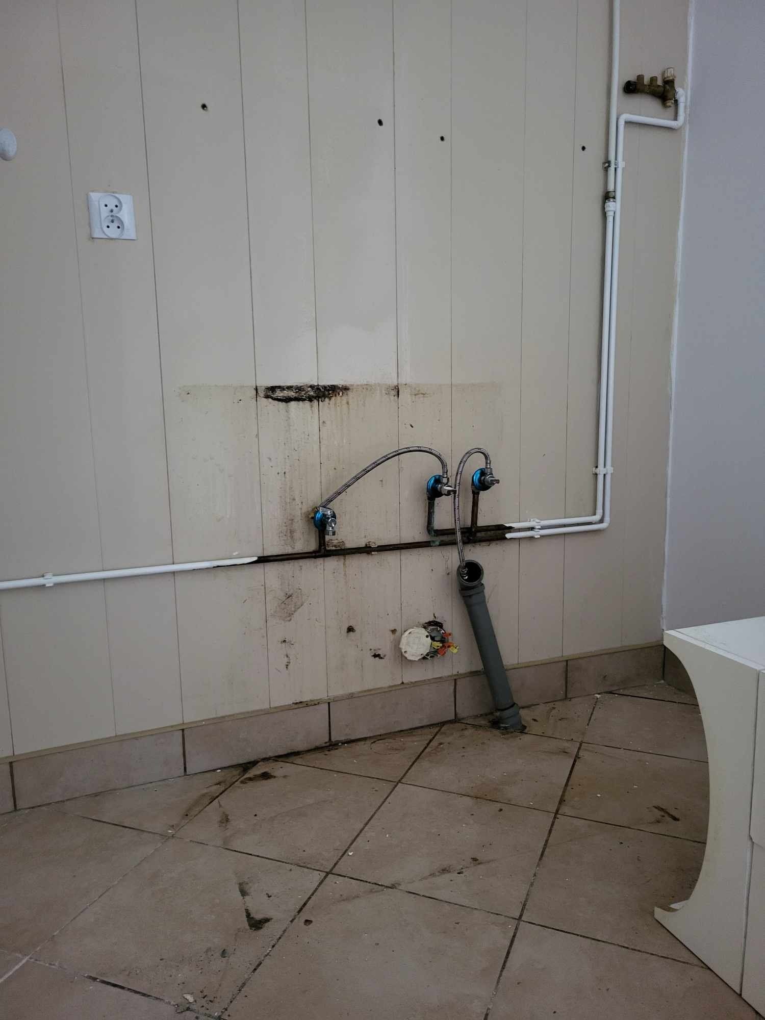 Ściana z panelami, widoczne rury, zawory, przyłącza kanalizacyjne i elektryczne przed montażem umywalki, ślady po starej instalacji i zabrudzenia na ścianie i podłodze.