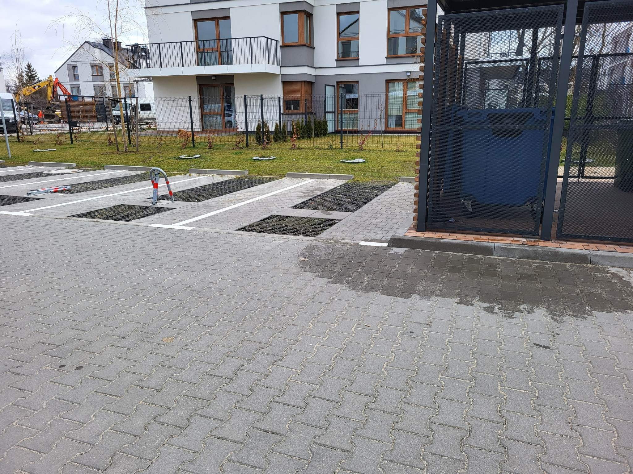 Nowoczesny parking z kostki brukowej przy apartamentowcu, widoczne miejsca parkingowe z ażurowymi płytami, trawnik z młodymi drzewkami i kontener na śmieci w metalowej klatce.