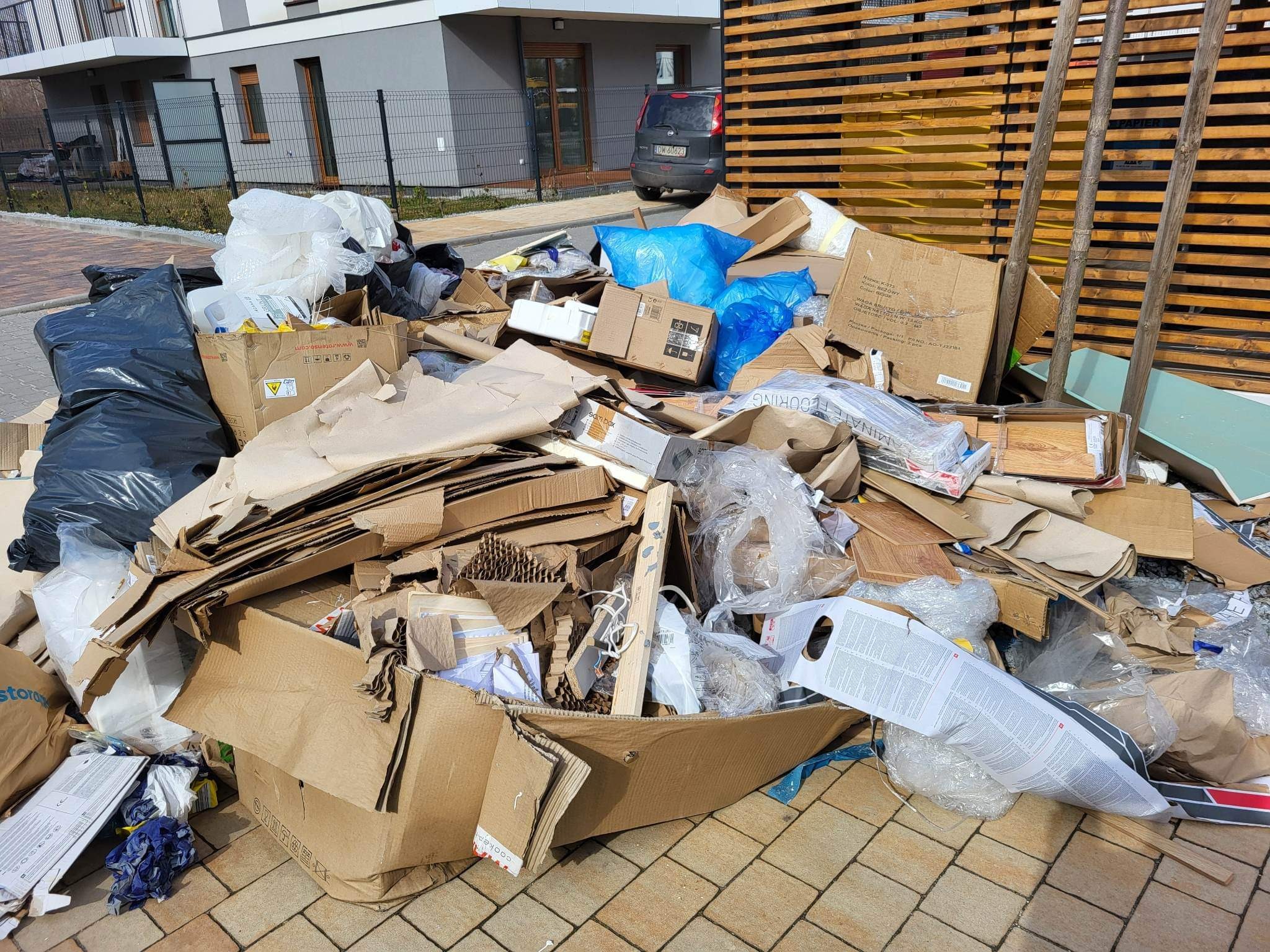 Stos kartonów, folii i innych odpadów po remoncie zgromadzony przed budynkiem mieszkalnym we Wrocławiu. Widoczne fragmenty opakowań po panelach podłogowych i innych materiałach budowlanych.