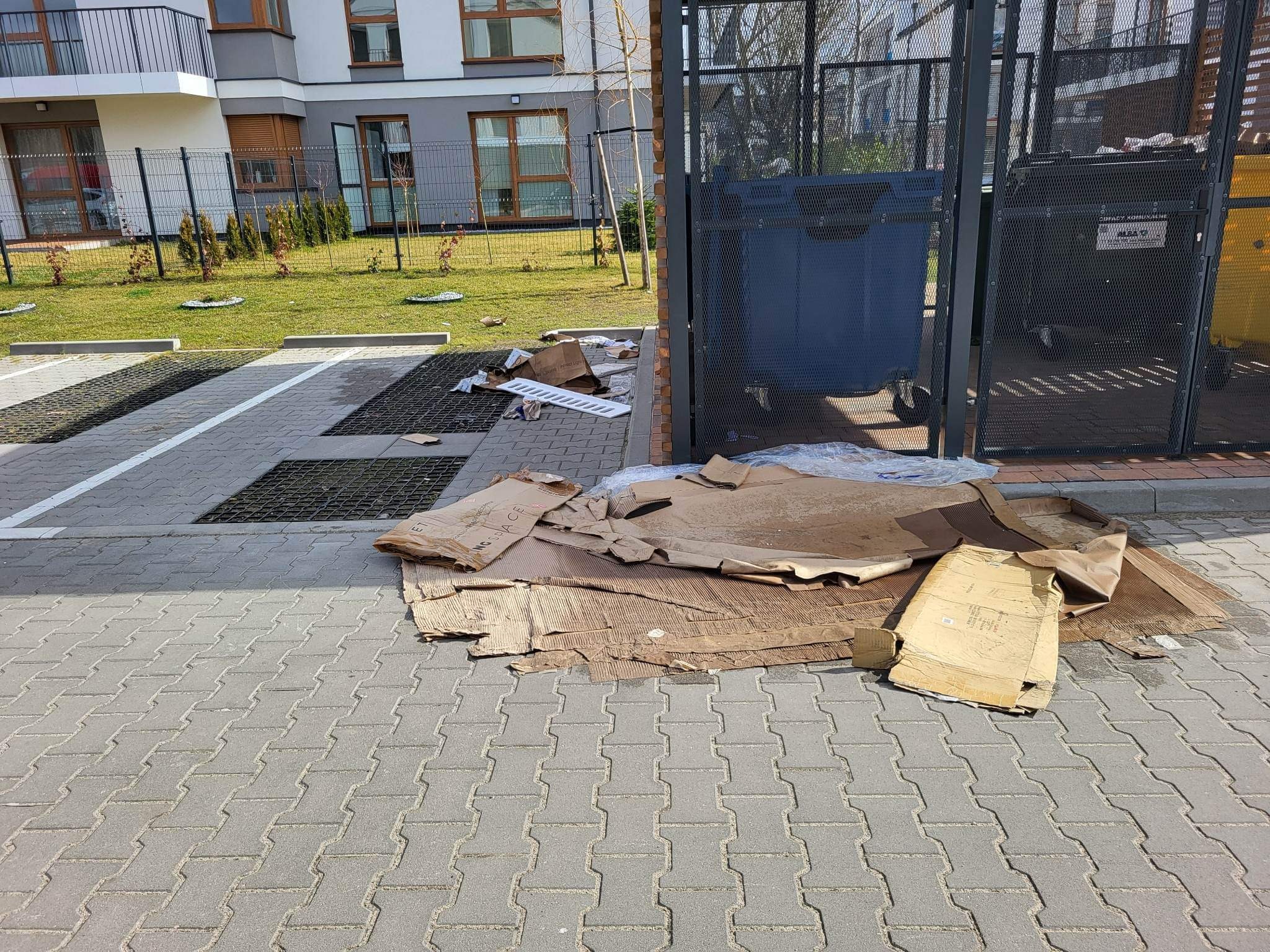 Stos rozłożonych kartonów i innych śmieci leży obok metalowej wiaty śmietnikowej na kostce brukowej, w tle widoczny budynek mieszkalny z zielonym trawnikiem.