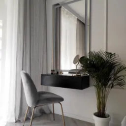 Elegancka toaletka z lustrem w srebrnej ramie, czarnym blatem i szarą welurową pufą na złotych nogach, uzupełniona o roślinę doniczkową i kosmetyki, na parkiecie w jodełkę.