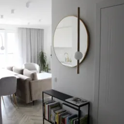 Nowoczesne wnętrze: parkiet w jodełkę, beżowa sofa, okrągłe lustro z lampą, czarny regał z książkami, białe ściany i drzwi.