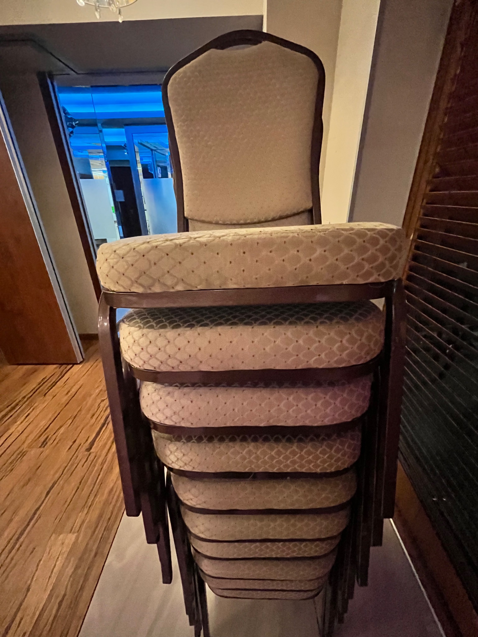 Stos krzeseł z tapicerowanymi siedziskami i oparciami, ustawionych jedno na drugim, widocznych na tle drewnianej podłogi i ściany z żaluzjami.