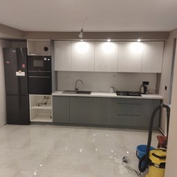 Budownictwo - Nowoczesna kuchnia w Nowodworze po remoncie, z szarymi szafkami dolnymi i białymi górnymi, czarnym sprzętem AGD i błyszczącą podłogą. Widoczny odkurzacz i wiadro, sugerujące sprzątanie po remoncie.