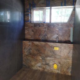 Budownictwo - Łazienka w trakcie wykańczania: brązowe płytki imitujące kamień na ścianach, ciemne, połyskujące płytki na sąsiedniej ścianie, drewnopodobne panele na podłodze, okno zabezpieczone taśmą malarską.