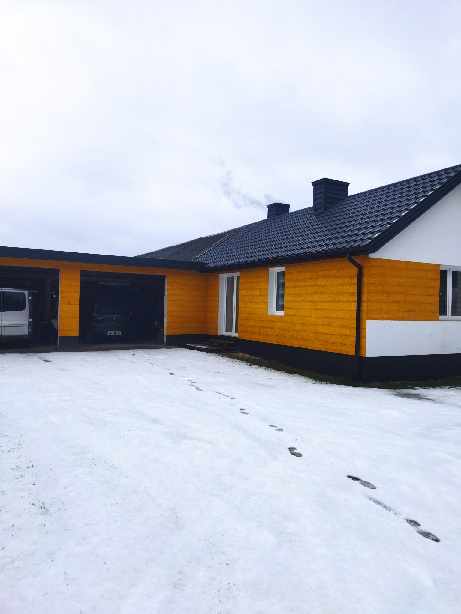 Żółta elewacja domu z czarnym dachem i dwoma garażami w zimowej scenerii, widoczne ślady butów na śniegu prowadzące do domu.