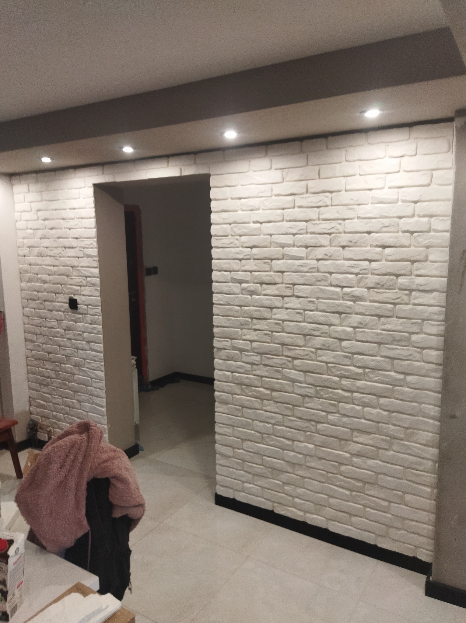 Ściana wykończona jasną cegłą dekoracyjną z widocznym wejściem do pokoju i fragmentem mebli z ubraniami w tle.