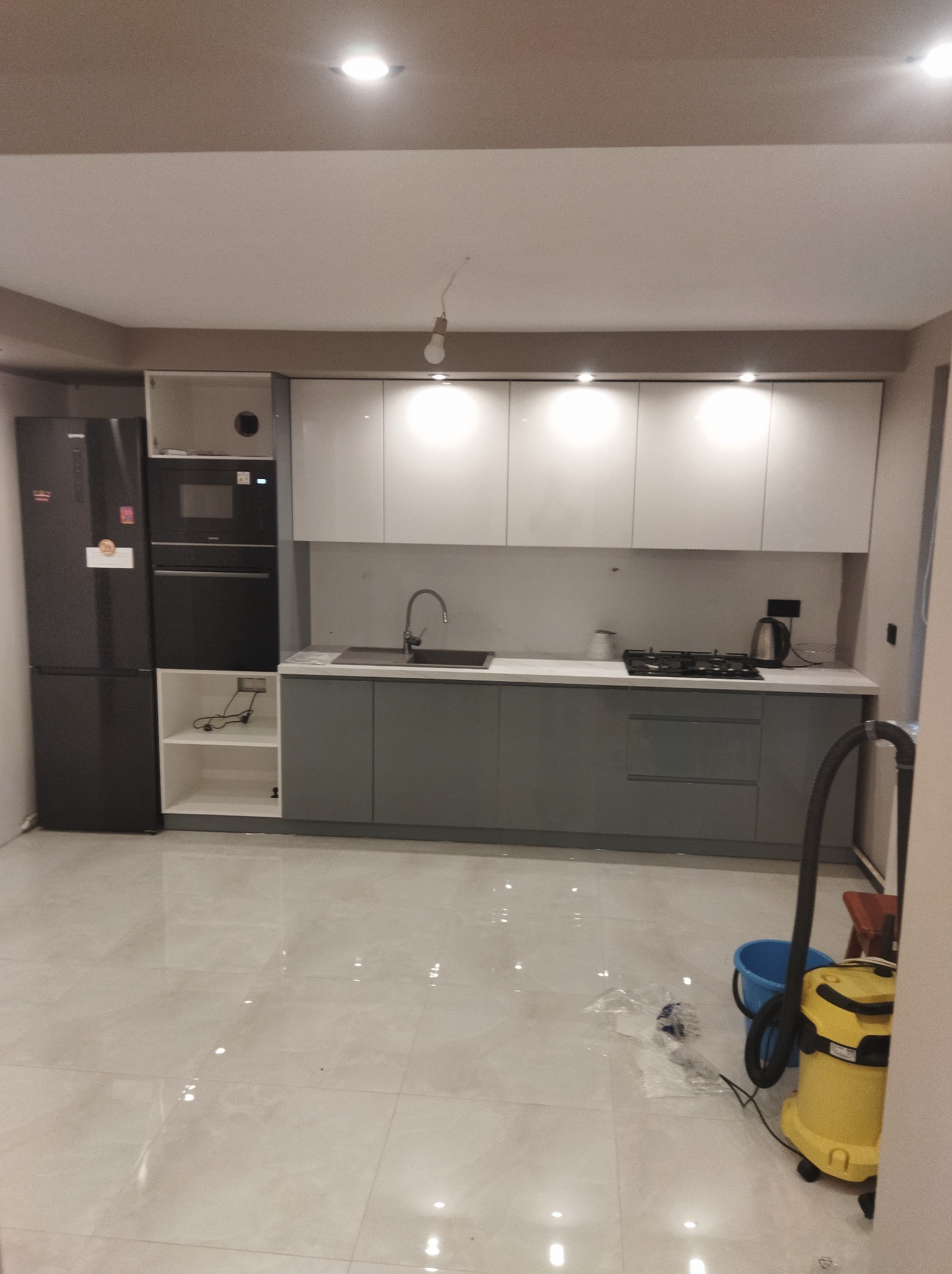 Nowoczesna kuchnia w Nowodworze po remoncie, z szarymi szafkami dolnymi i białymi górnymi, czarnym sprzętem AGD i błyszczącą podłogą. Widoczny odkurzacz i wiadro, sugerujące sprzątanie po remoncie.