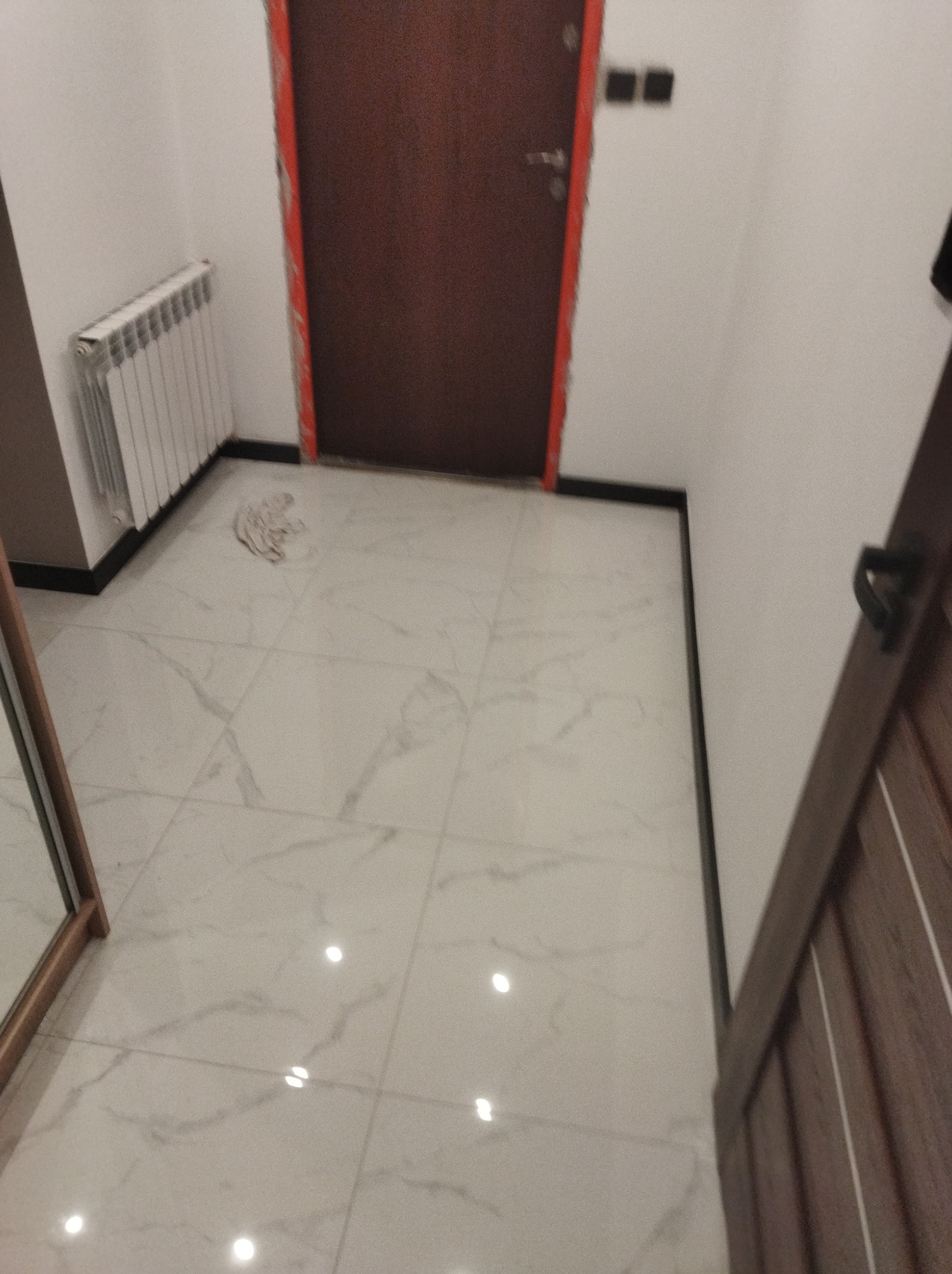 Wykończone wnętrze: białe marmurowe płytki podłogowe, nowy grzejnik żeberkowy, ciemnobrązowe drzwi z czarnymi listwami przypodłogowymi.