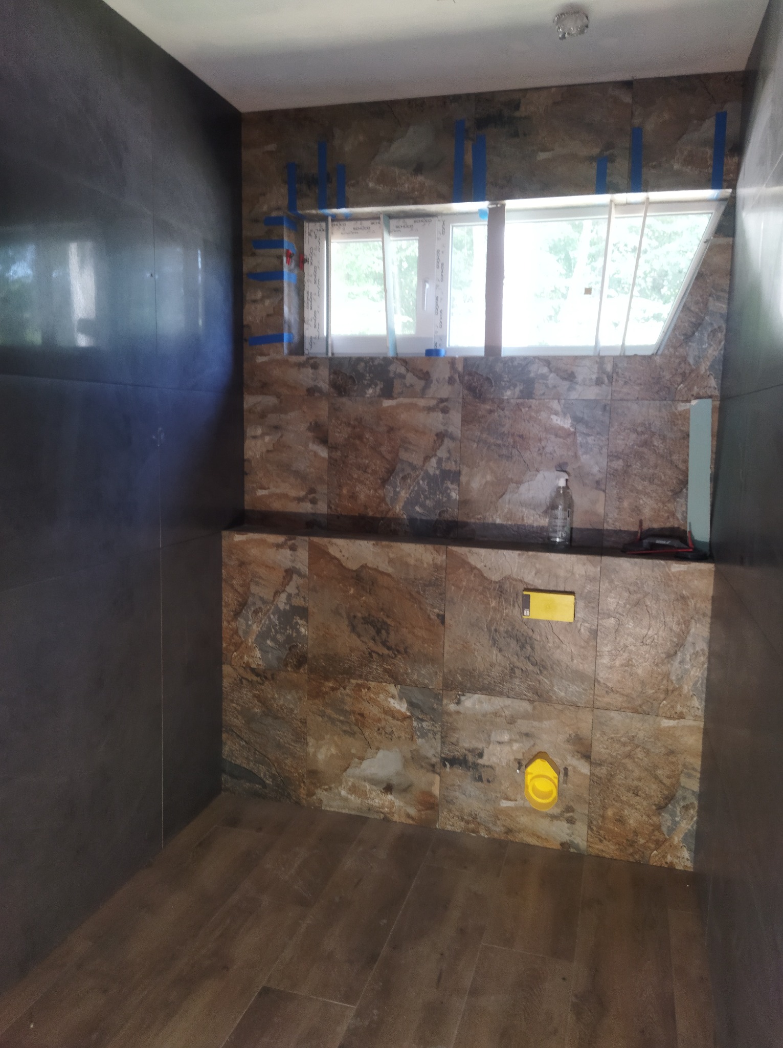 Łazienka w trakcie wykańczania: brązowe płytki imitujące kamień na ścianach, ciemne, połyskujące płytki na sąsiedniej ścianie, drewnopodobne panele na podłodze, okno zabezpieczone taśmą malarską.