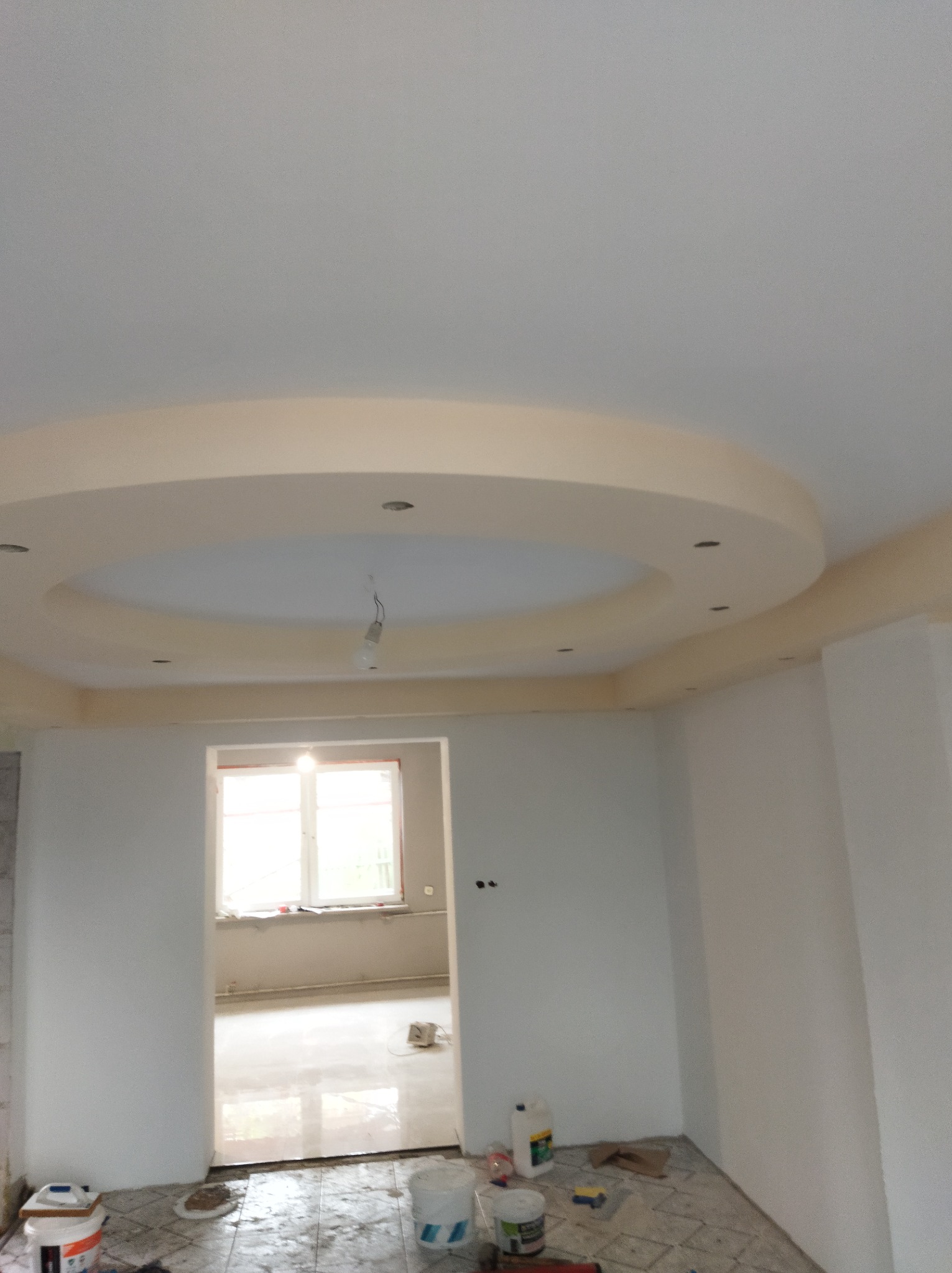 Wnętrze pokoju po remoncie z dekoracyjnym okrągłym podwieszanym sufitem z punktowym oświetleniem, widoczne ślady prac wykończeniowych na podłodze i wiaderka z farbą.
