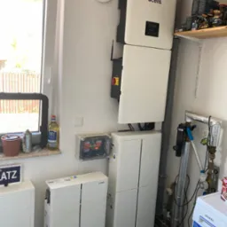 Domowa instalacja fotowoltaiczna z inwerterem i akumulatorami marki Qcells, widoczna skrzynka elektryczna i elementy instalacji hydraulicznej oraz pralka Siemens w tle.