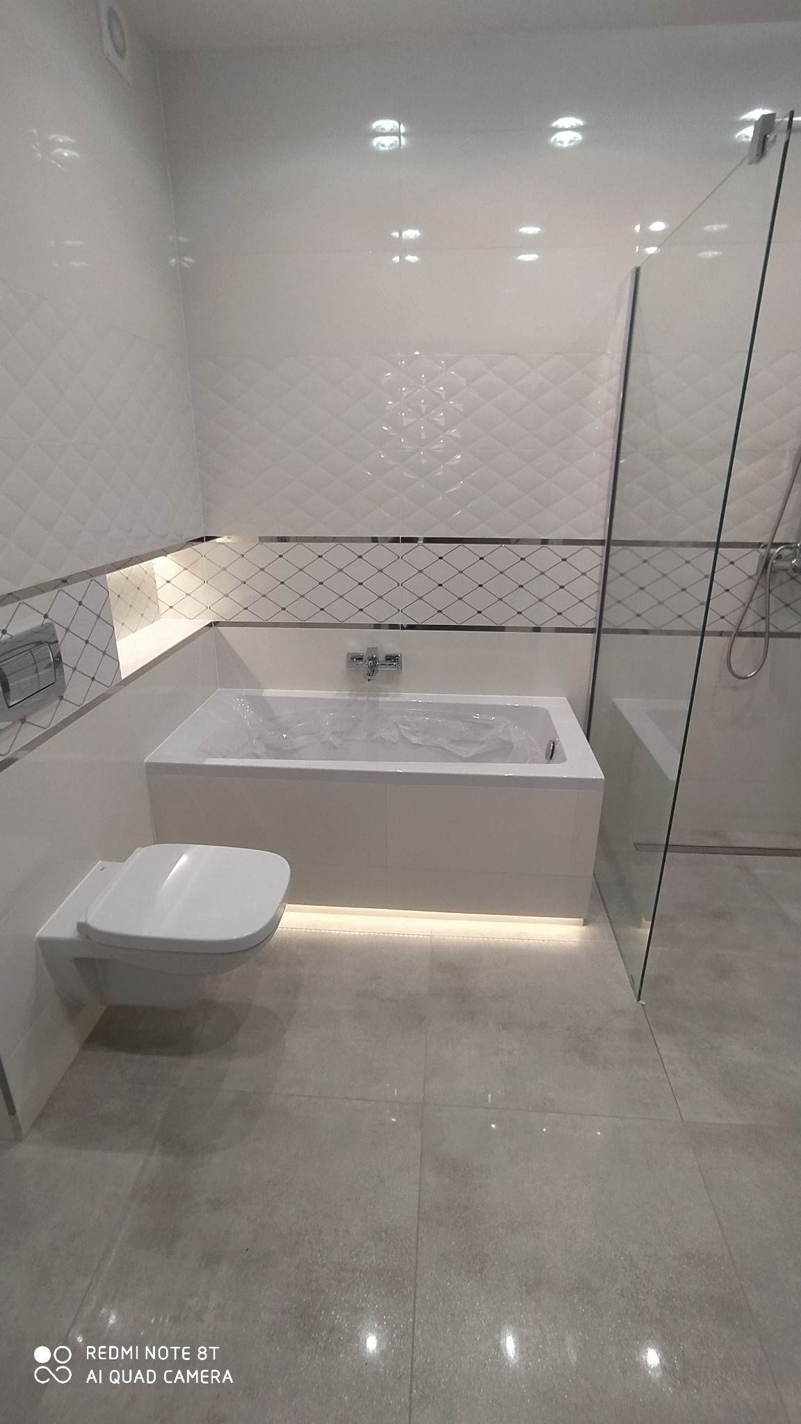 Nowoczesna łazienka z białą armaturą, podświetloną wanną, szklaną kabiną prysznicową i geometrycznymi płytkami na ścianach. Wanna zabezpieczona folią ochronną.