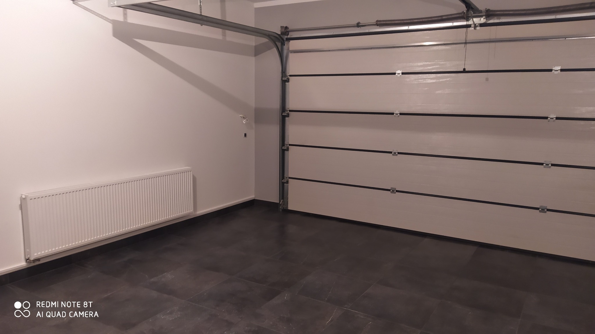 Wnętrze garażu po remoncie: ściany pomalowane na jasny kolor, ciemna, błyszcząca podłoga z płytek, biały grzejnik na ścianie, segmentowa brama garażowa. Widoczne elementy konstrukcyjne bramy...