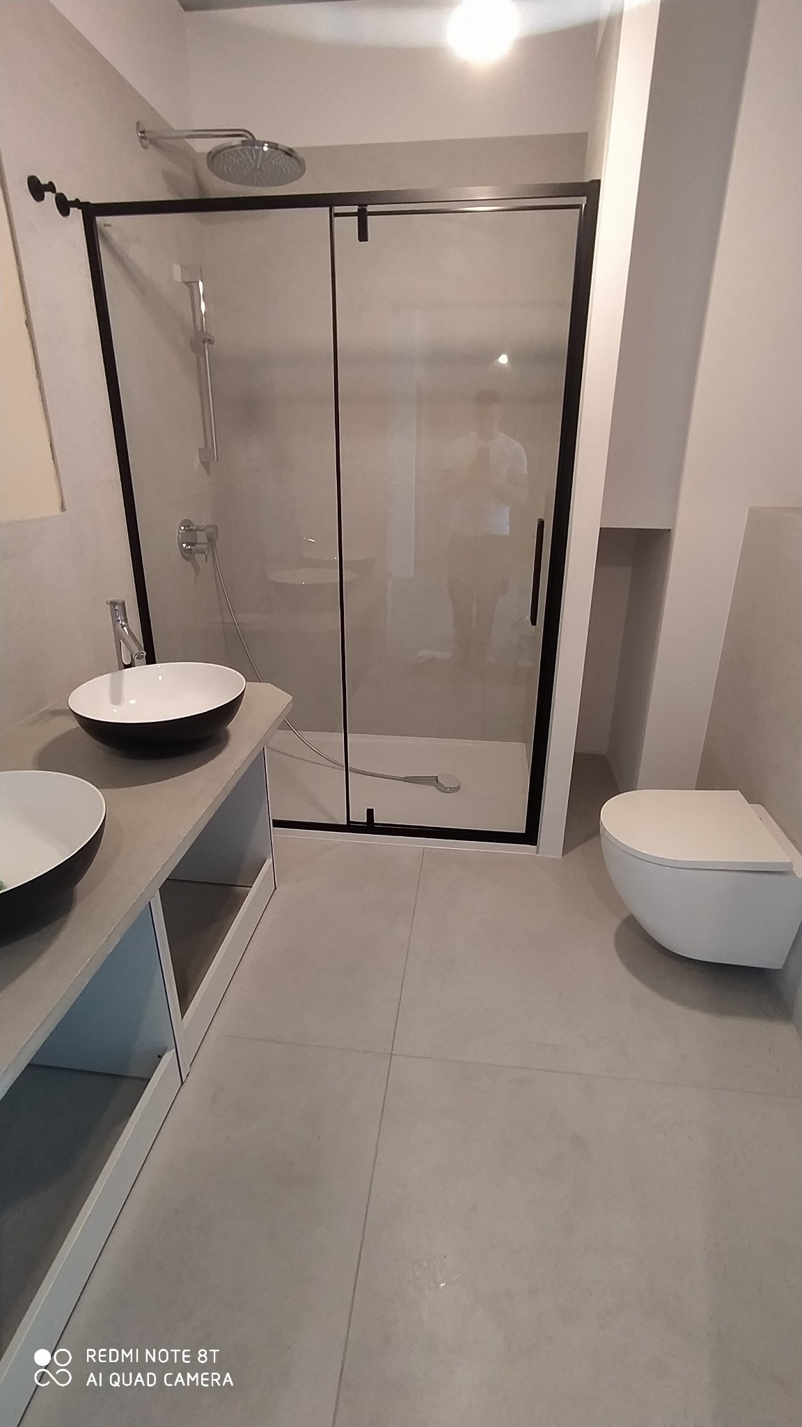 Nowoczesna łazienka z kabiną prysznicową z czarną ramą, umywalkami nablatowymi w kolorze czarno-białym oraz podwieszaną miską WC w minimalistycznym stylu.