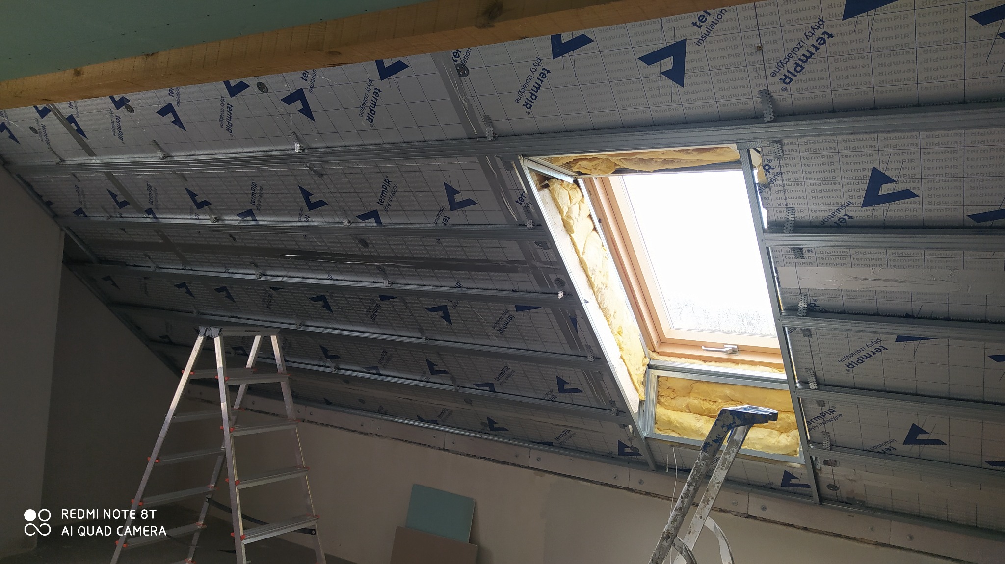 Poddasze w trakcie ocieplania, widoczne płyty izolacyjne TermPIR z folią aluminiową, metalowa konstrukcja stelażu pod płyty kartonowo-gipsowe oraz okno dachowe z izolacją z wełny mineralnej.