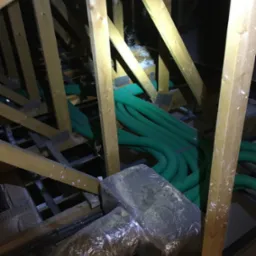 Instalacja wentylacyjna z zielonymi przewodami w przestrzeni poddasza, widoczne drewniane elementy konstrukcji dachu i zaizolowane kanały wentylacyjne.