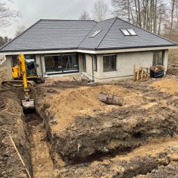 WAGREX bis Sebastian Piątkowski - Żółta koparka wykopuje wąski rów pod fundamenty domu jednorodzinnego z szarą dachówką, widoczne palety z drewnem i stos czarnej folii.